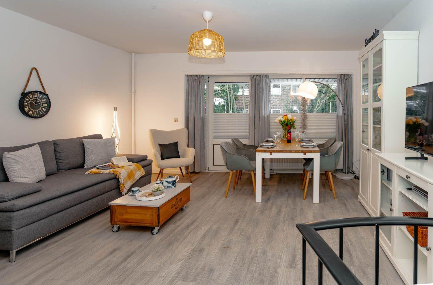 Wohn- und Essbereich mit Sofa, Tisch und Stühlen. Holzboden und Fenster mit Vorhängen.