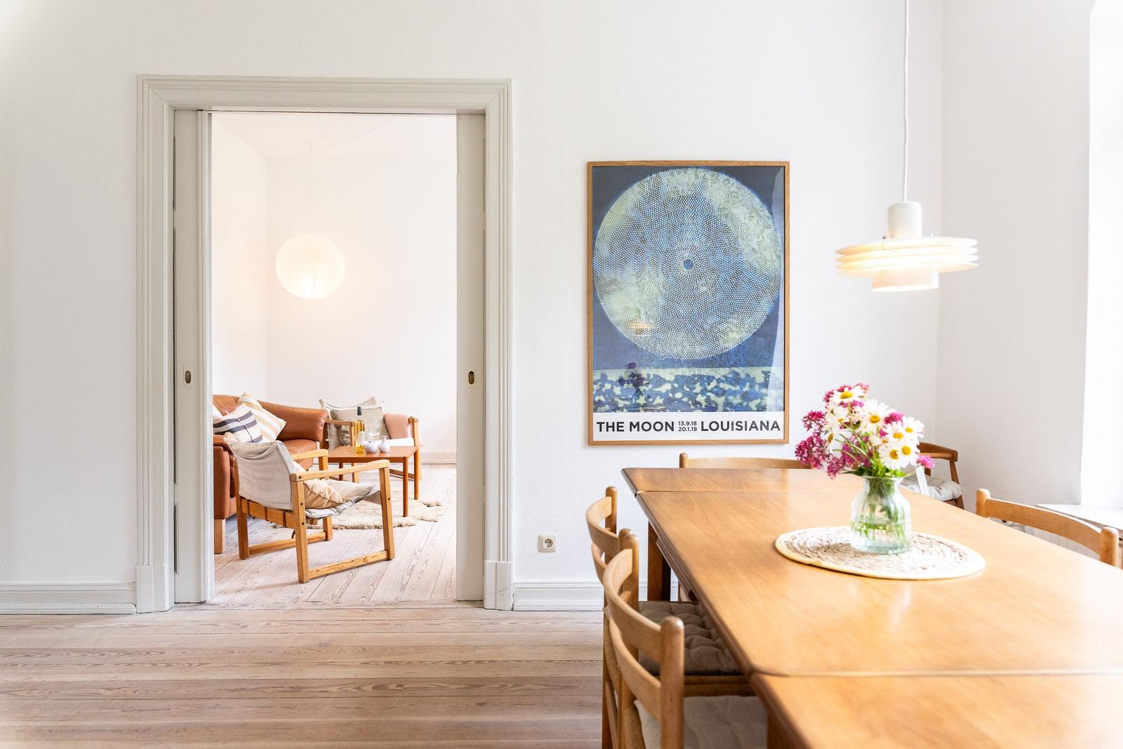Esszimmer mit Holztisch und Stühlen. Wandbild und Blumen auf Tisch. Durchgang zum Wohnzimmer.