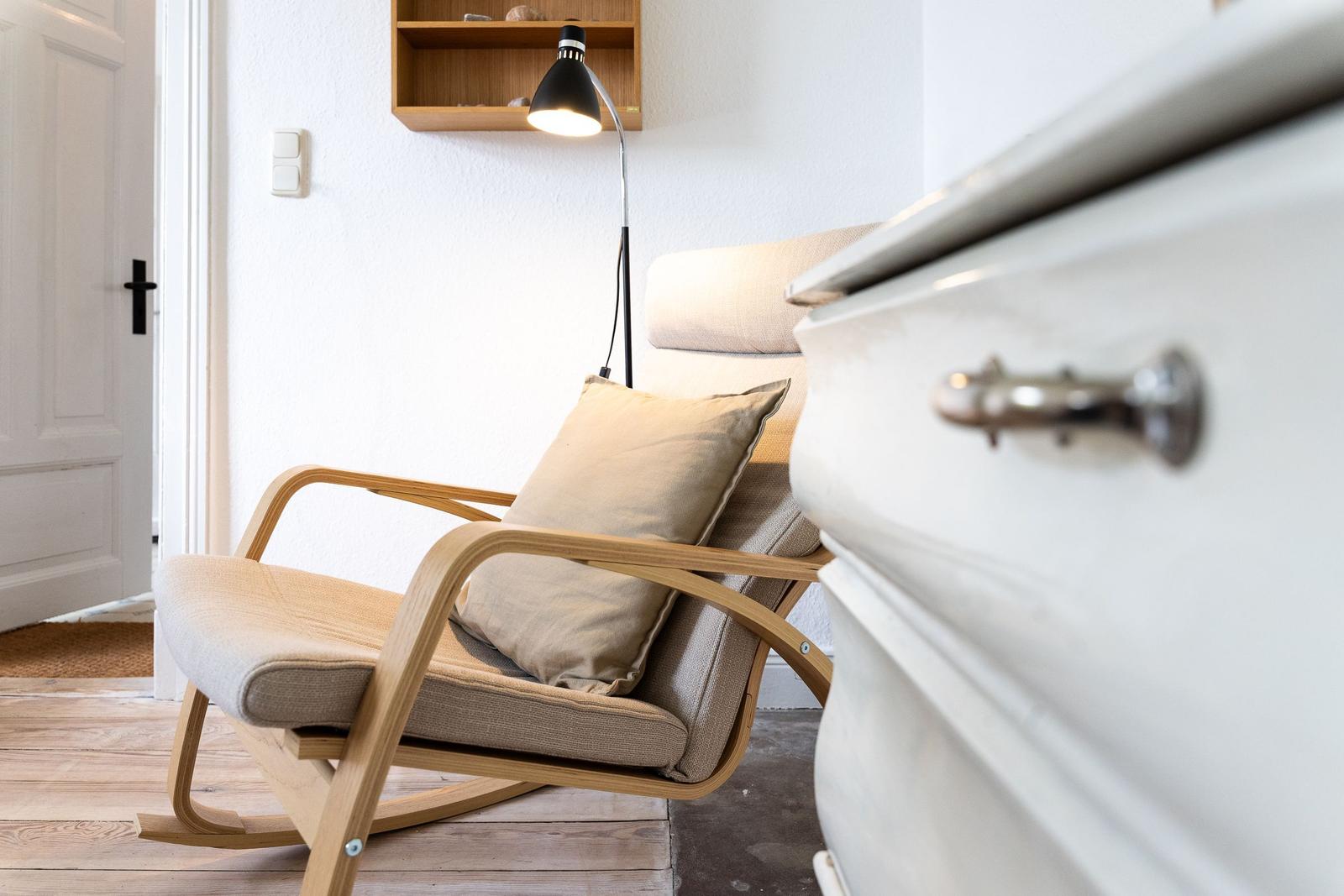 Ein Holzschaukelstuhl mit Kissen steht neben einem weißen Schrank. Eine Stehlampe beleuchtet den Raum.