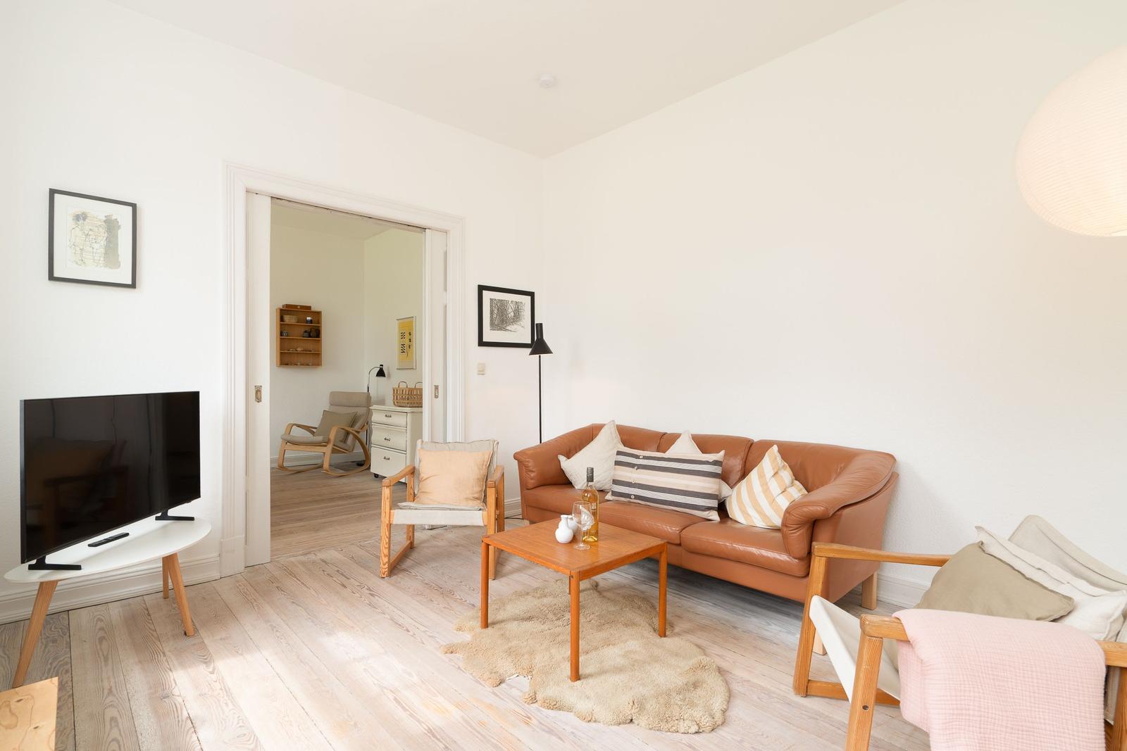 Wohnzimmer mit braunem Sofa, Holzfußboden und Fernseher. Durchgang zu einem anderen Raum.