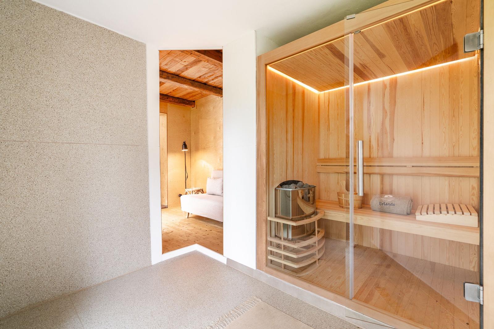 Sauna mit Holzverkleidung und Glaswand. Im Hintergrund ist ein Schlafzimmer mit Holzboden und Bett sichtbar.