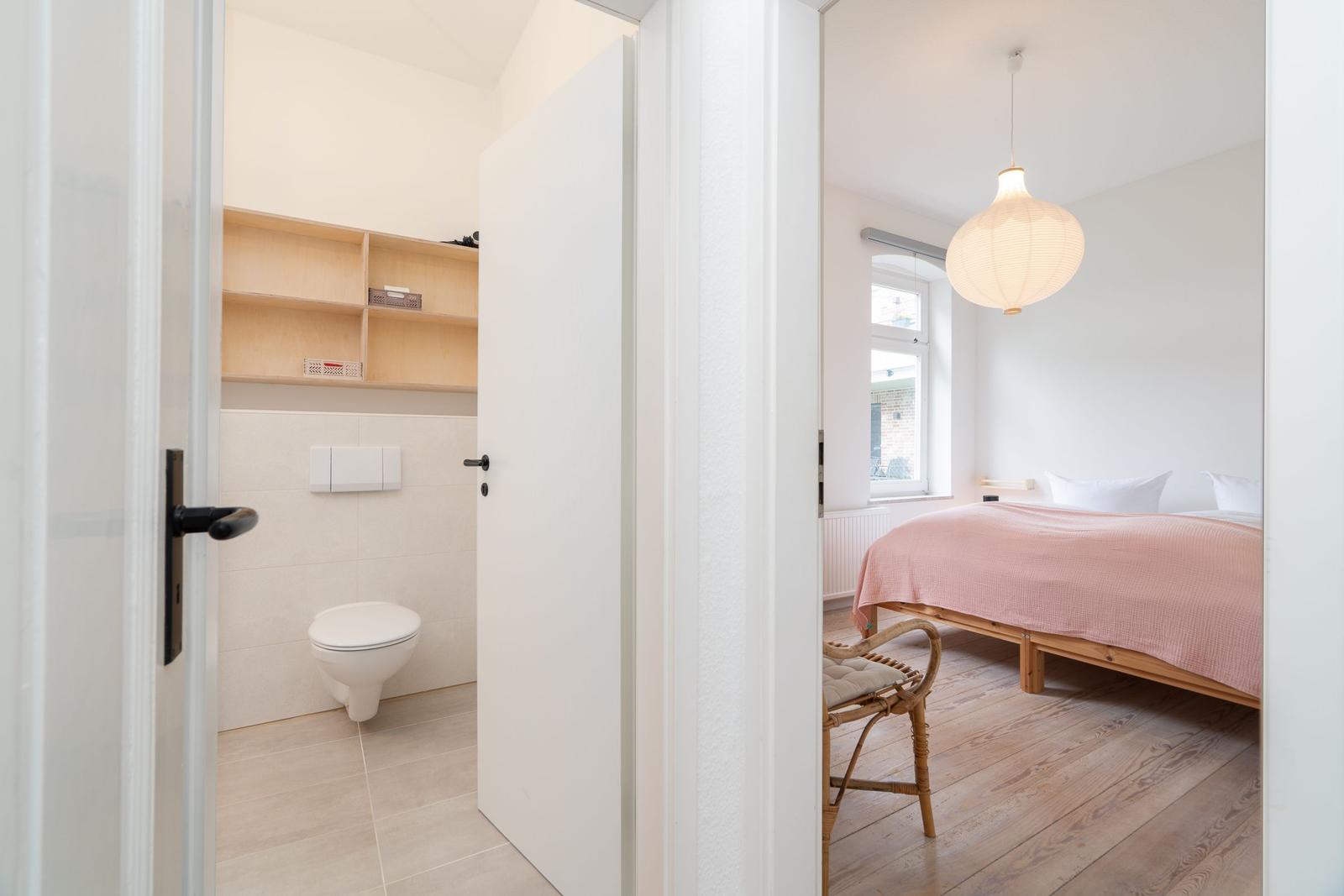 Schlafzimmer mit Bett und Fenster. Nebenraum mit Toilette und Regal.