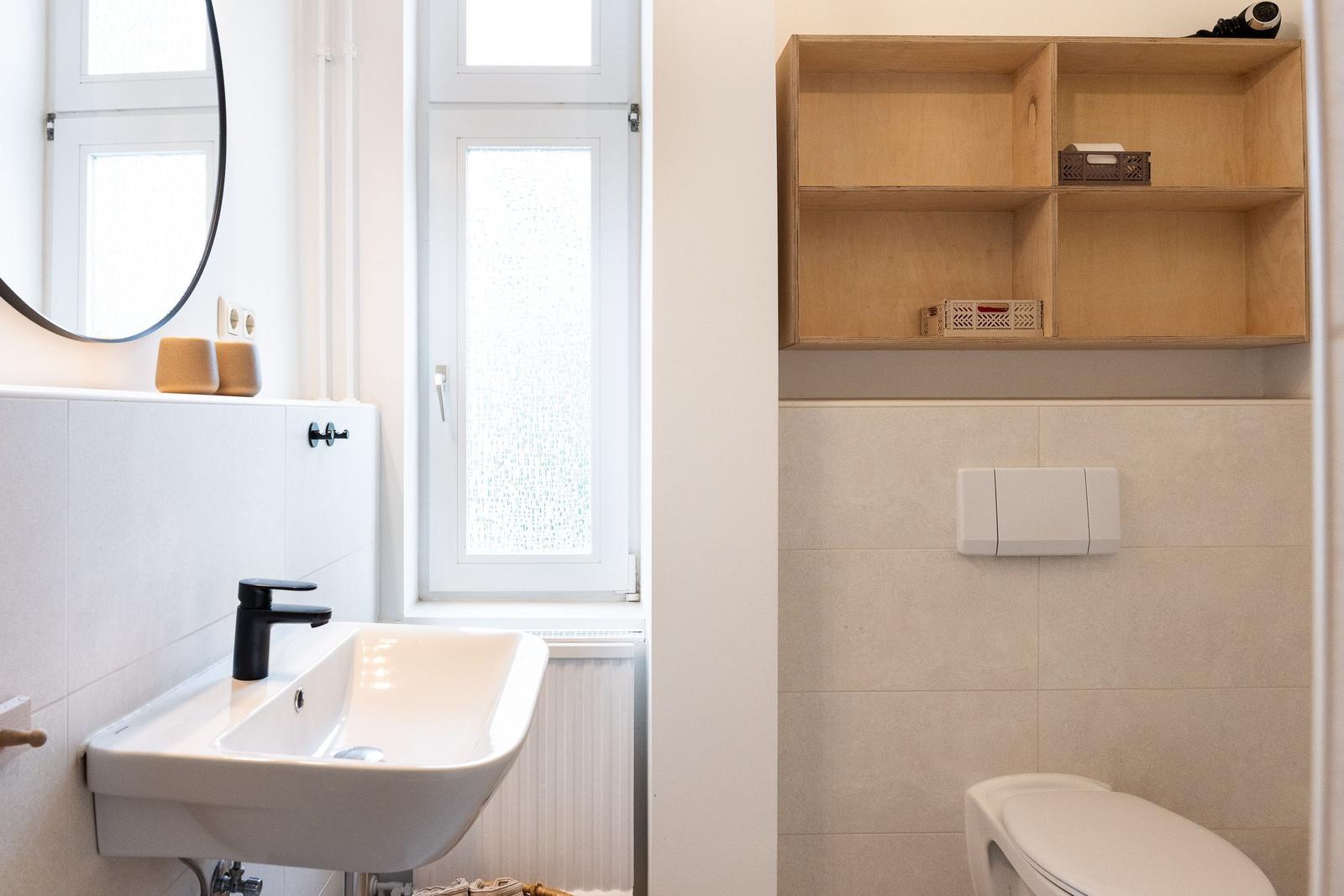 Badezimmer mit Waschbecken, Spiegel, Fenster und Toilette. Holzregal an der Wand.