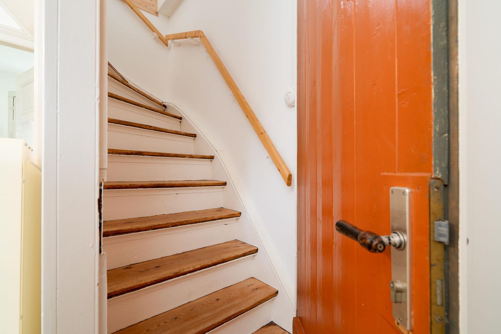 Helle Treppe mit Holzstufen und Geländer neben einer orangefarbenen Tür.