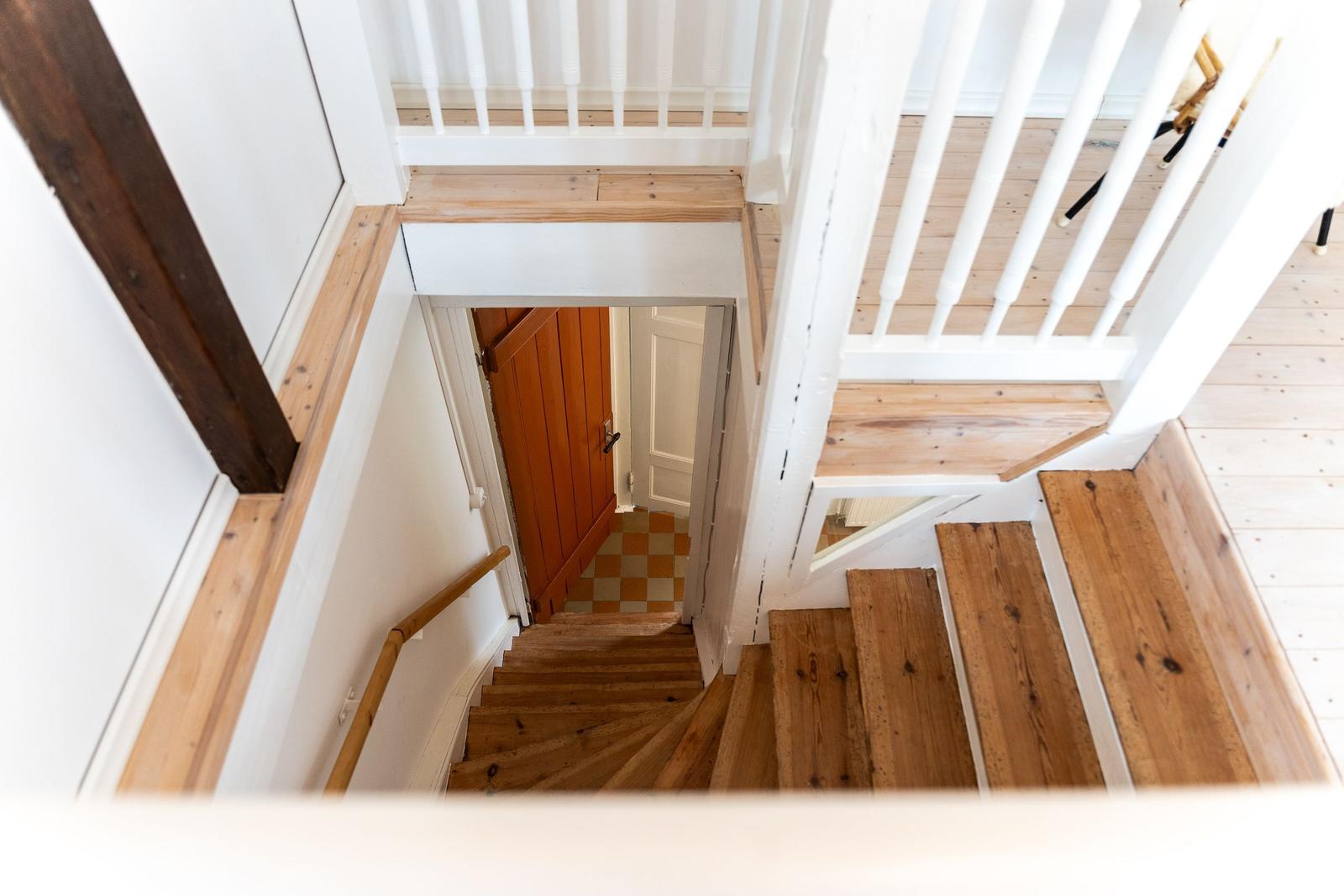 Holzgetreppe mit weißem Geländer führt nach unten. Tür und Kachelboden sind sichtbar.