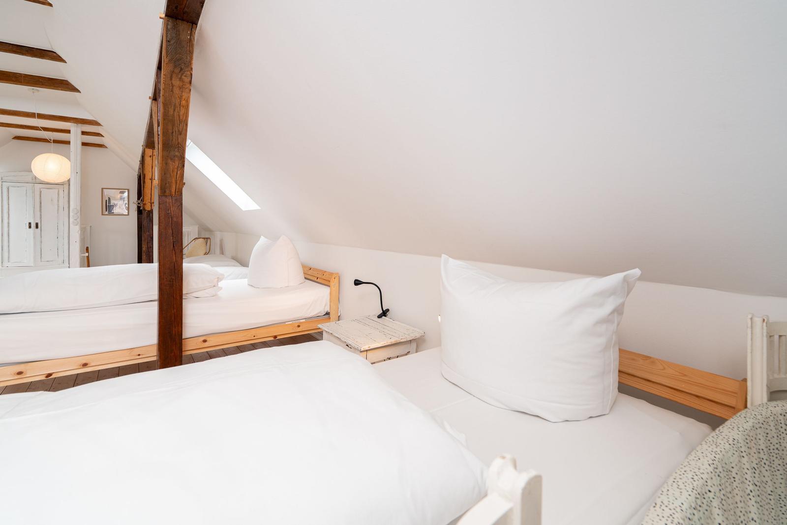 Zimmer mit zwei Betten, Dachschräge und Holzbalken. Fenster und Nachttisch sichtbar.