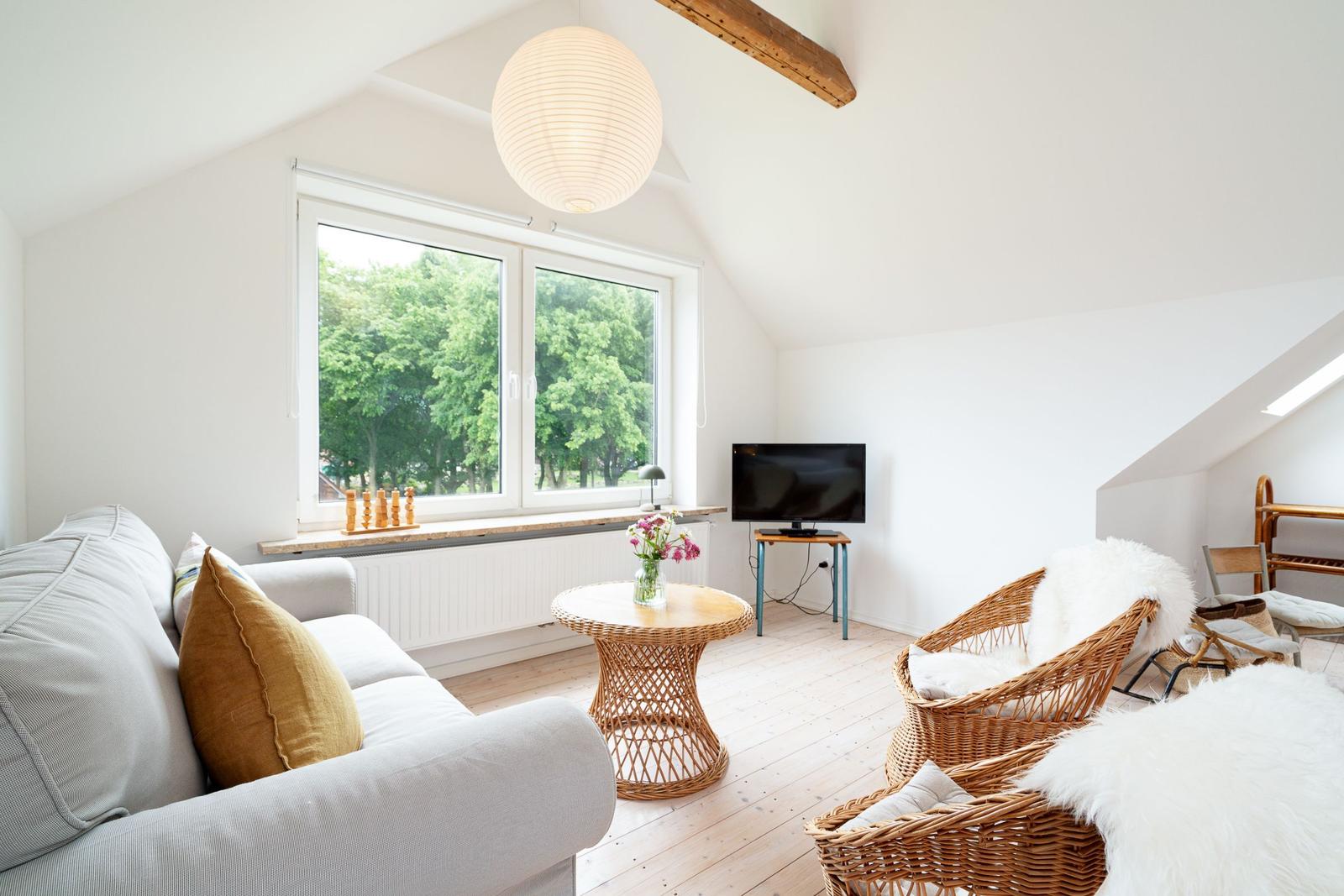 Wohnzimmer mit Sofa, Rattanstühlen und TV. Fenster mit Blick auf Bäume.