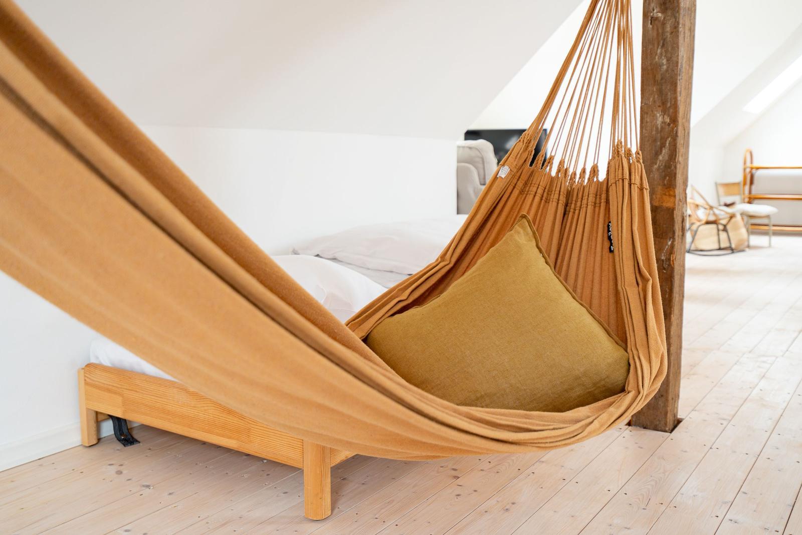 Hängemattensitz mit Kissen neben Bett und Holzbalken im Dachzimmer.