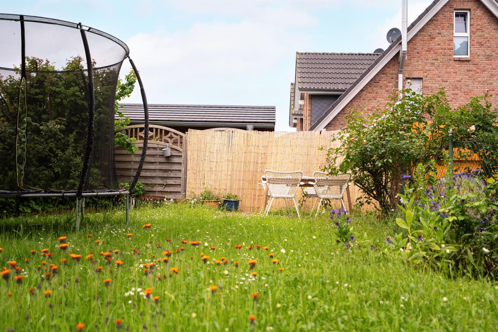 Hinterhof mit Trampolin, Gartenmöbeln und Blumen. Holzzaun und Nachbarhaus im Hintergrund.