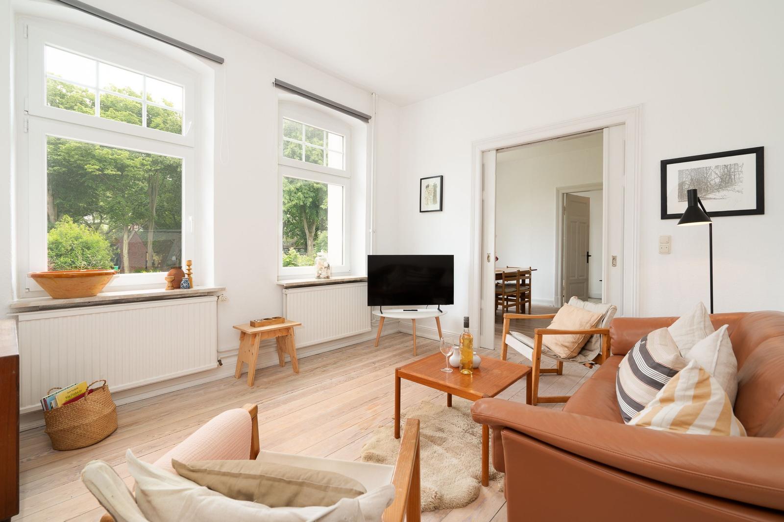 Wohnzimmer mit braunem Sofa, Holztisch und großen Fenstern. Blick auf grüne Bäume.