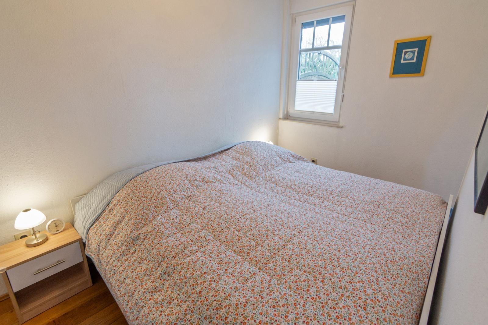 Schlafzimmer mit Bett, Nachttisch und Fenster. Wandbild und Lampe sind sichtbar.
