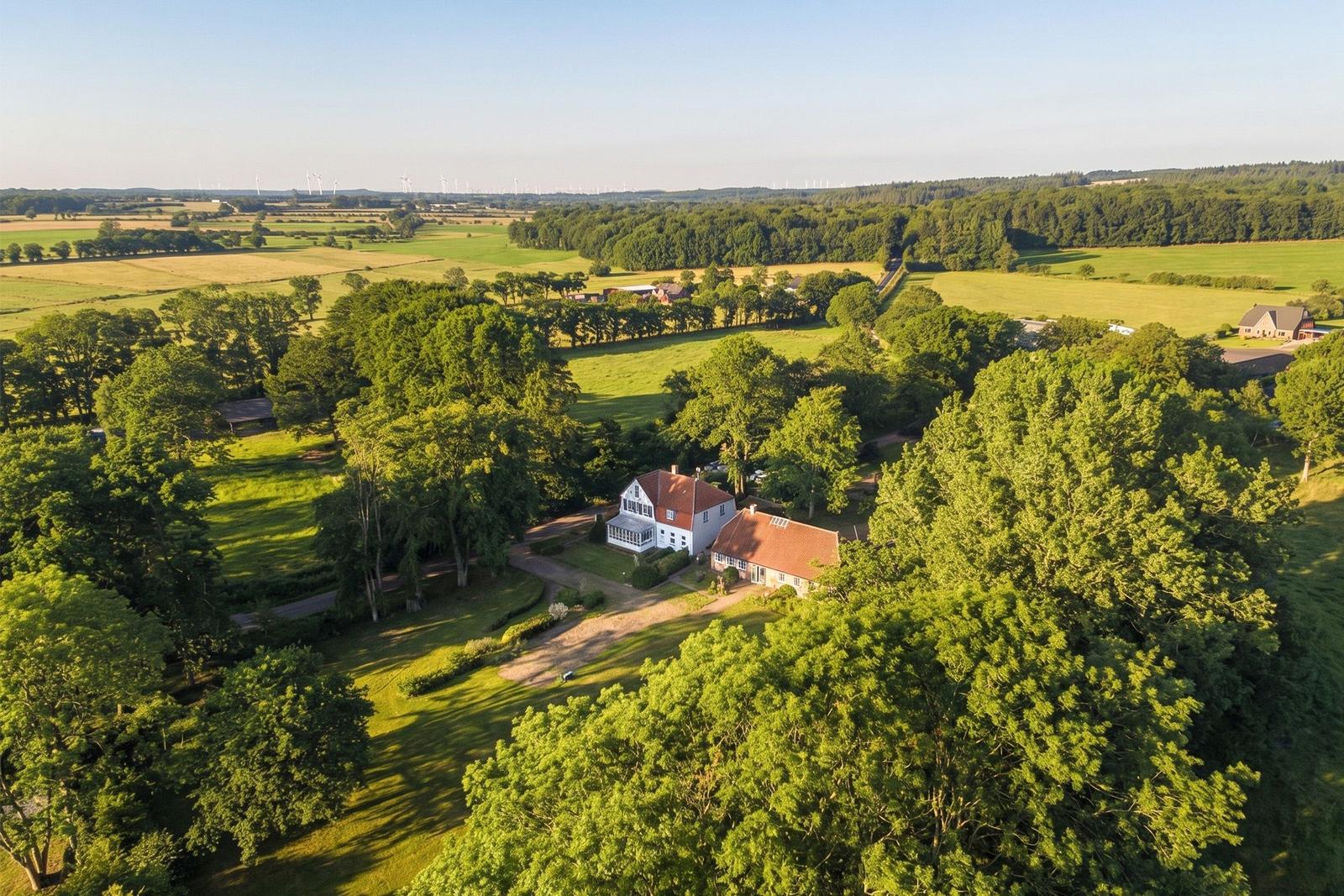 House with red roof, surrounded by green meadows and trees. Wind turbines are visible in the background.