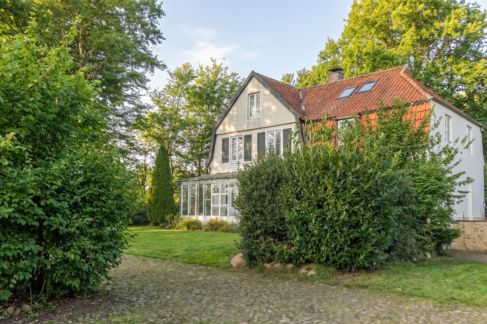 House with red roof, green garden, and gravel path. Surrounded by trees and shrubs.