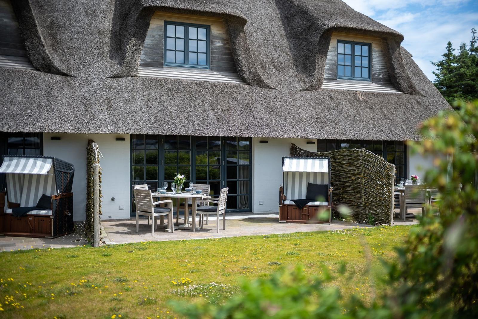 Haus mit Strohdach, Terrasse mit Tisch, Stühlen und Strandkörben.