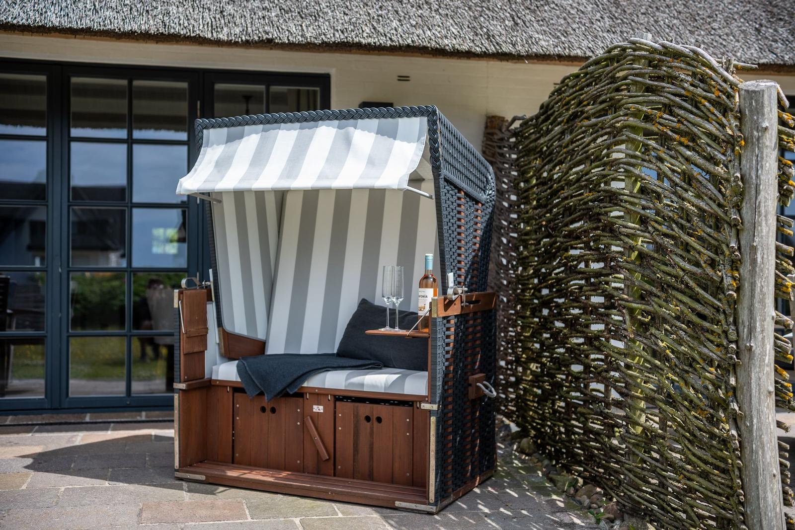 Strandkorb mit Streifen, Flasche und Gläsern vor einem Haus mit Strohdach und Holzzaun.