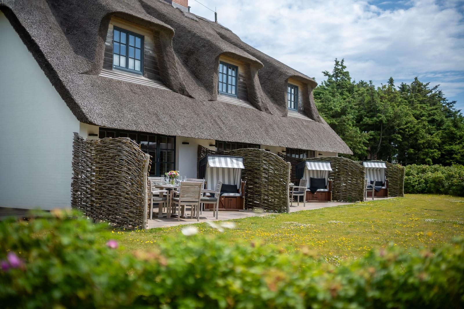 Haus mit Strohdach, Terrasse mit Tischen und Liegestühlen, Grasfläche mit Blumen.