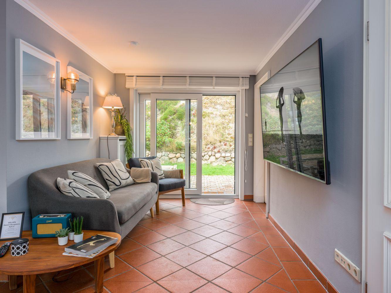 Wohnzimmer mit Sofa, Fernseher und Terrassentür. Terrasse mit Steinmauer und Grünflächen.