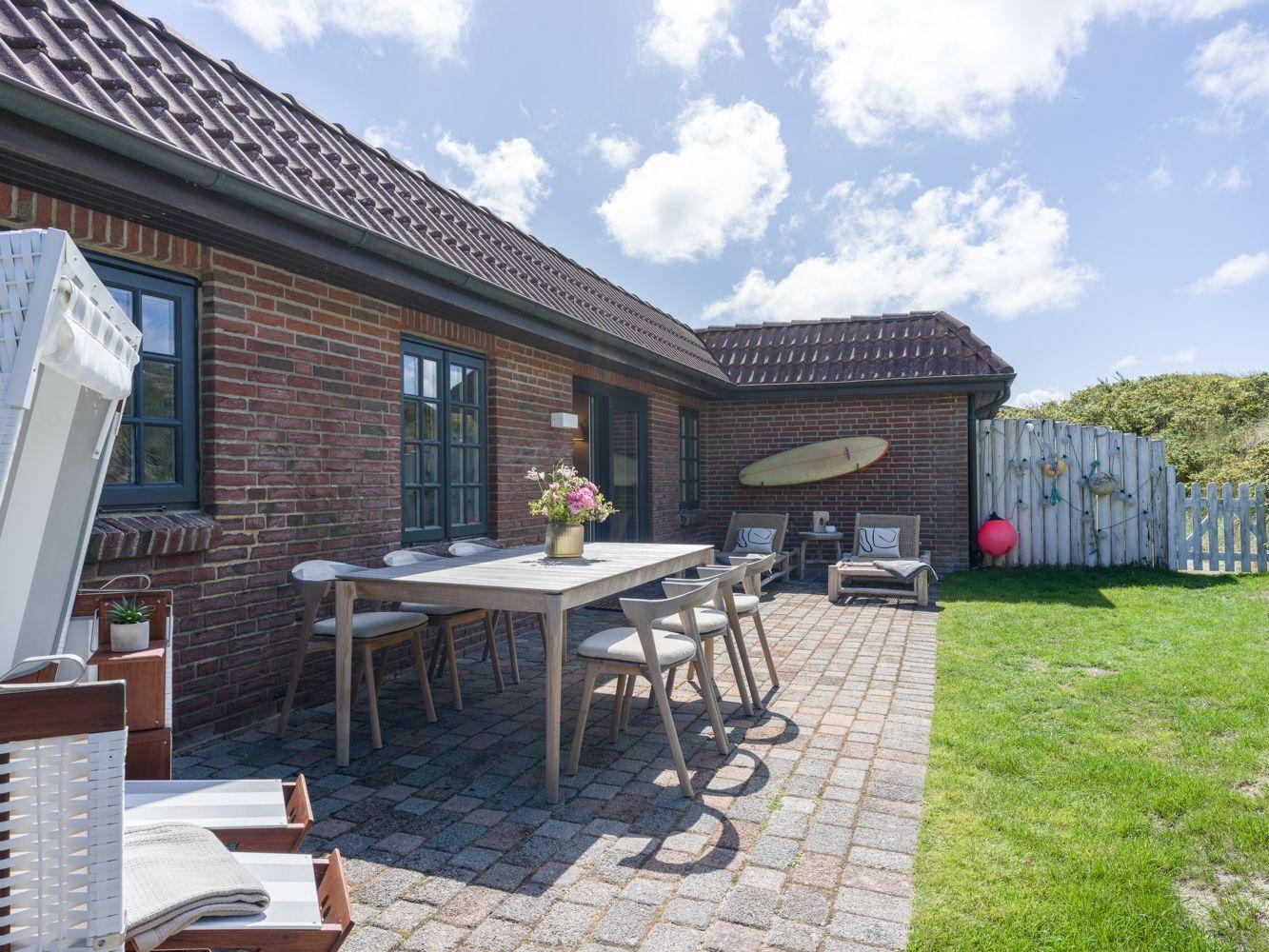 Roter Backsteinbau mit Terrasse, Tisch, Stühlen und Garten. Surfbrett an der Wand.