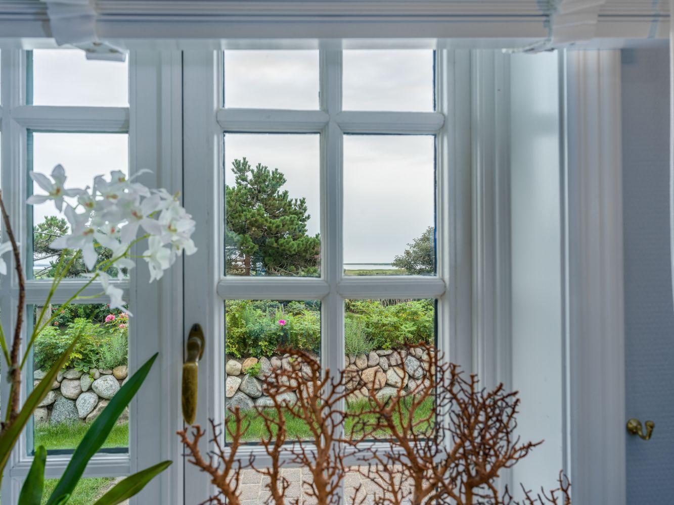 Durch das Fenster blickt man auf einen Garten mit Steinmauer und Bäumen.