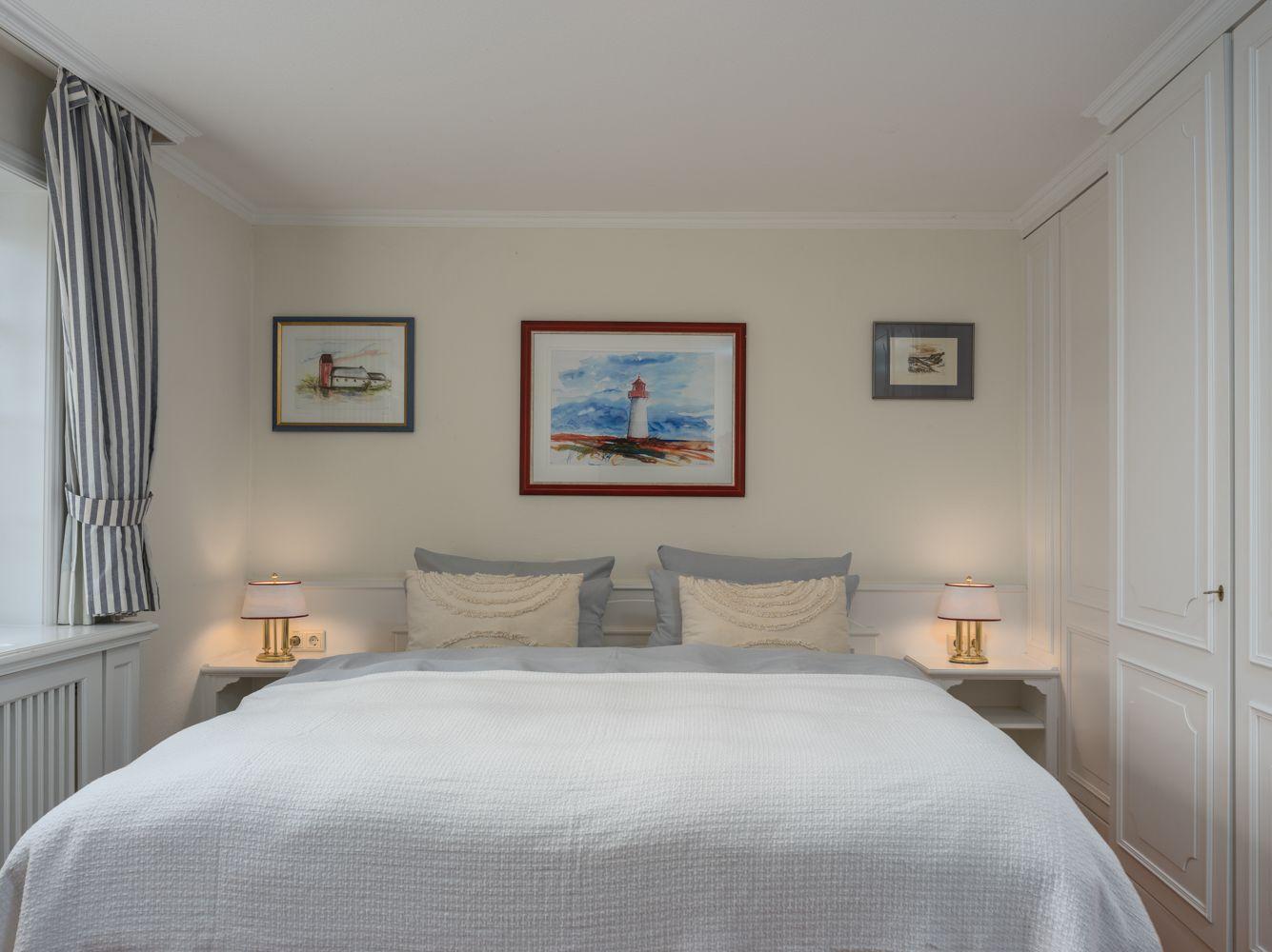Schlafzimmer mit Bett, Nachttischen und Wandbildern. Fenster mit Streifenvorhängen.