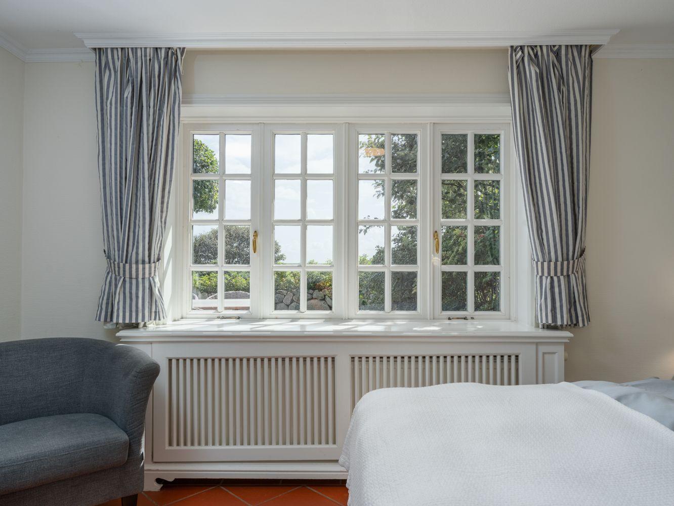Schlafzimmer mit großem Fenster, Bett und Sessel. Vorhänge und Heizkörper sind sichtbar.