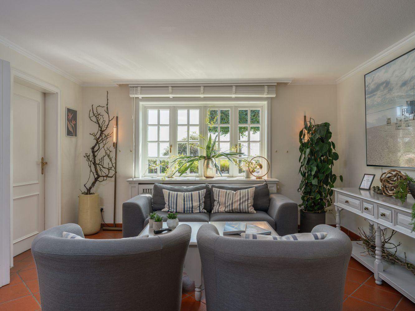 Wohnzimmer mit grauem Sofa, zwei Sesseln und großem Fenster. Terrakottaboden und Pflanzen.