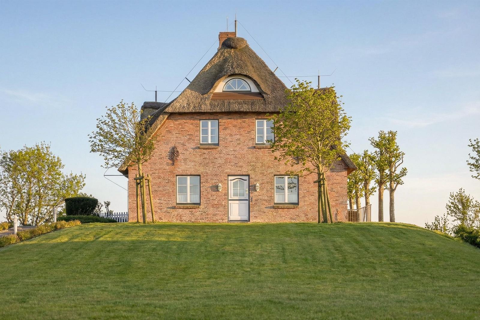 Rotes Backsteinhaus mit Strohdach auf grünem Hügel. Umgeben von Bäumen und klarer Luft.