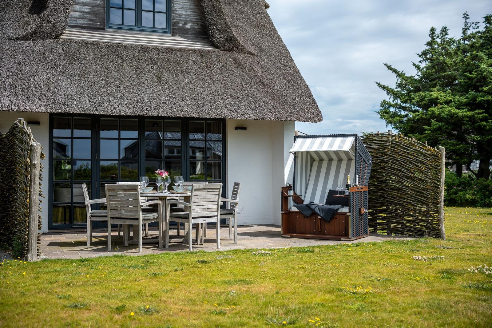 Haus mit Strohdach, Terrasse mit Tisch und Stühlen, Strandkorb und Grasfläche.