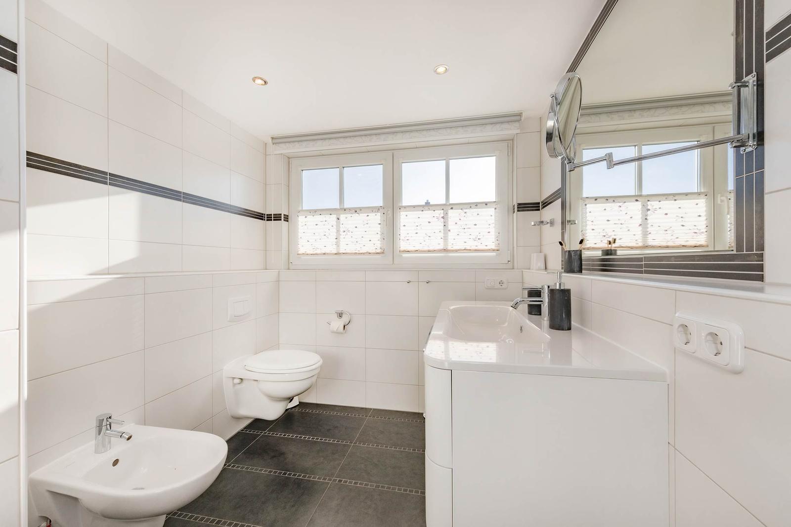 Badezimmer mit Waschbecken, Toilette und Spiegel. Fenster mit Jalousie. Fliesenboden.