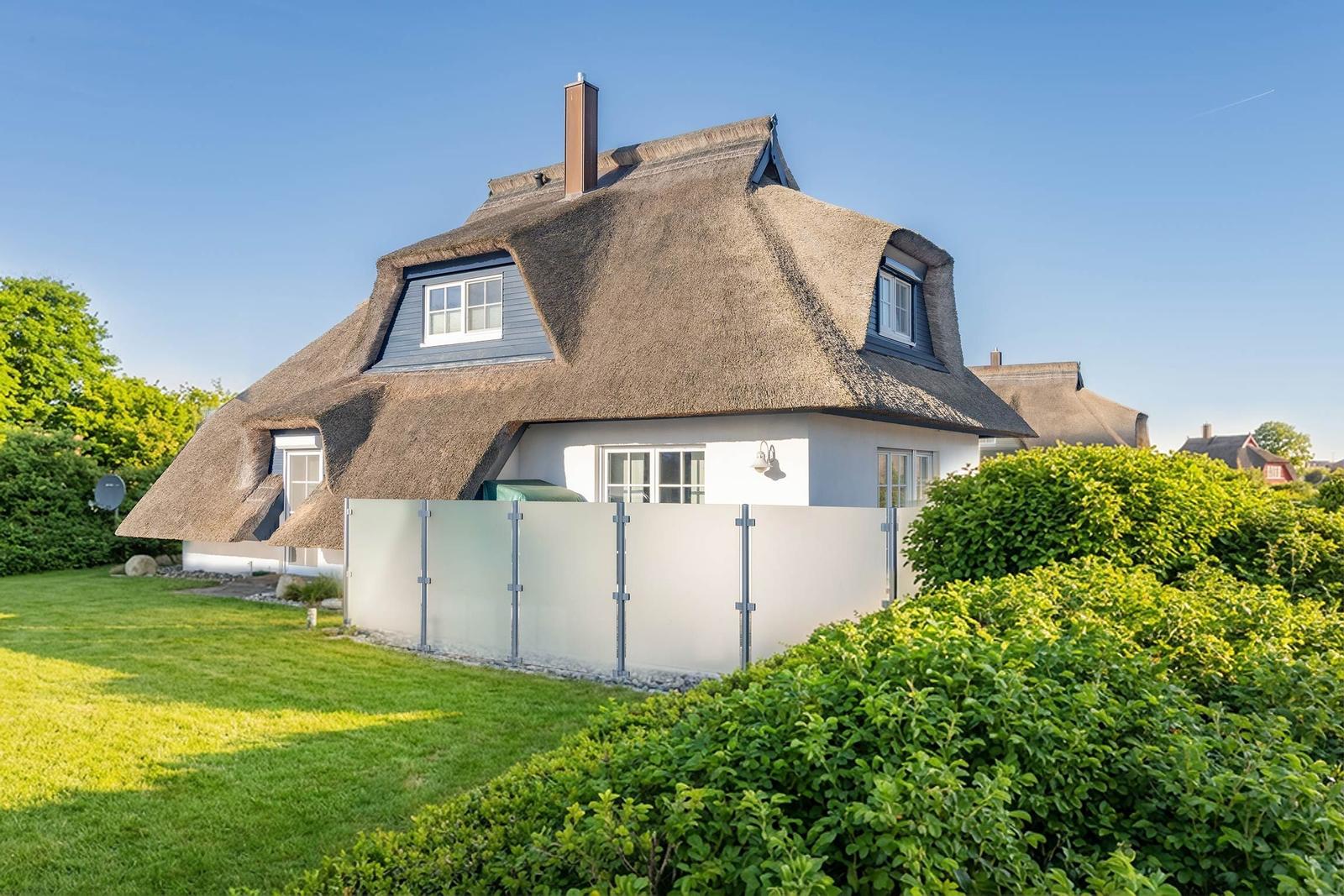 Dorfhaus mit Strohdach, weißer Fassade und Gartenzaun. Grünflächen und Sträucher umgeben das Gebäude.