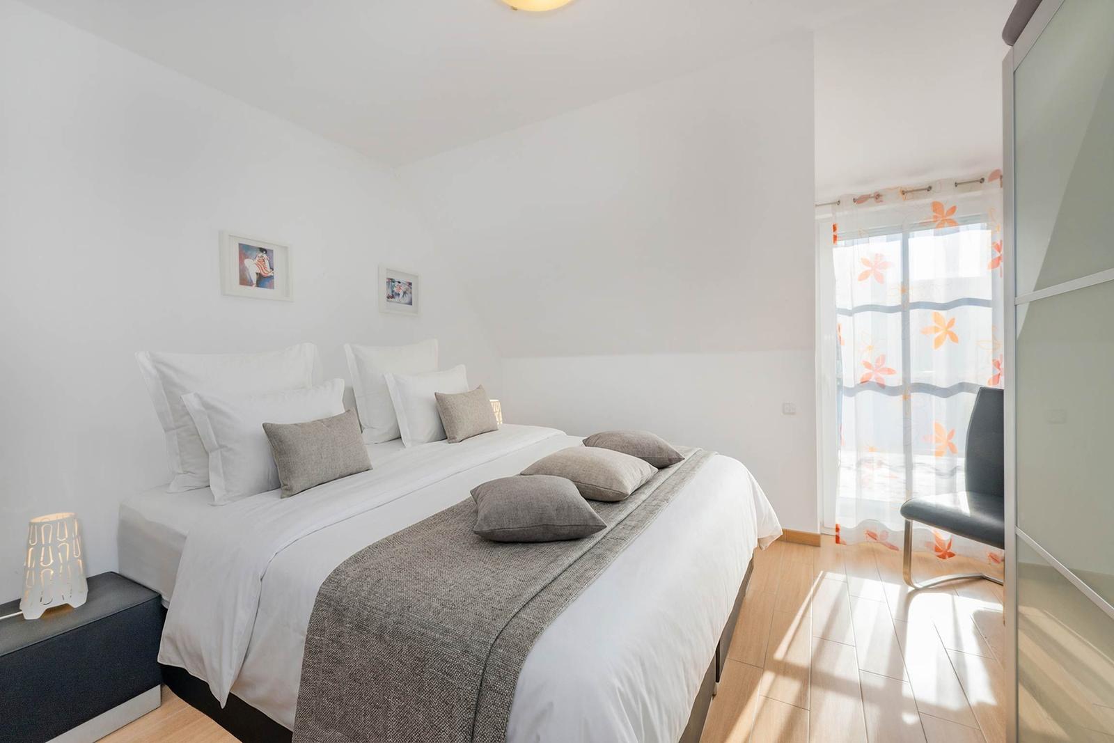 Schlafzimmer mit Doppelbett, weißen Wänden und Holzboden. Fenster mit floralen Vorhängen.