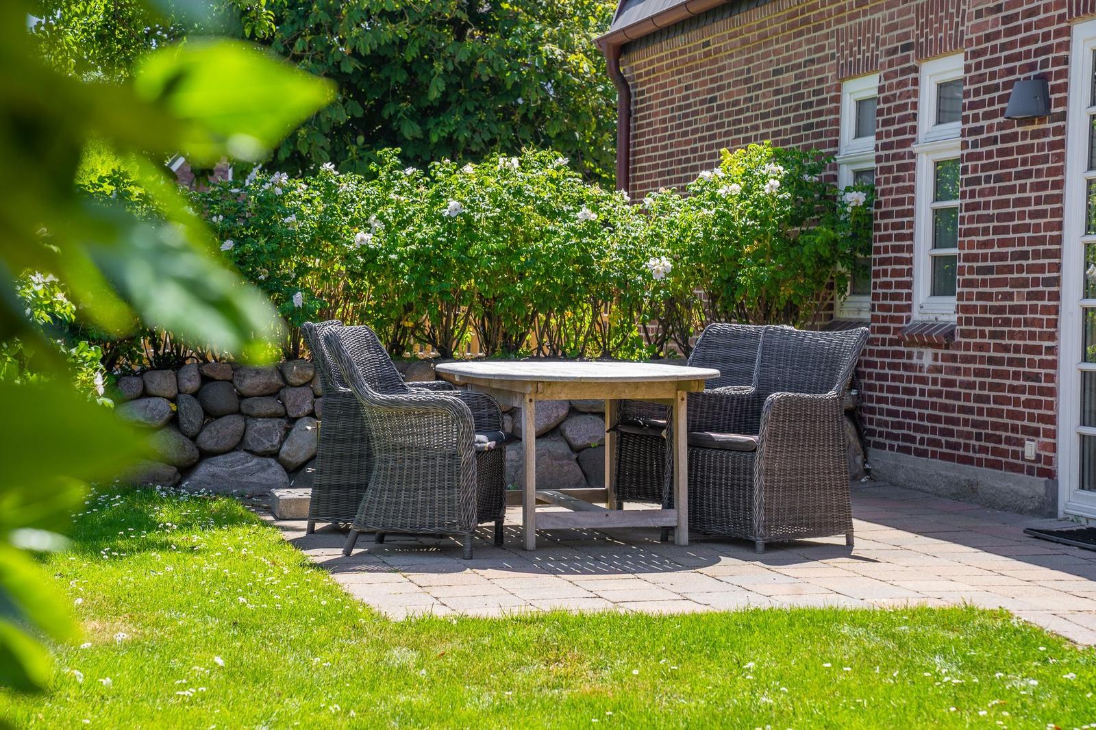 Patio with table and chairs in front of a brick house with flower hedge.