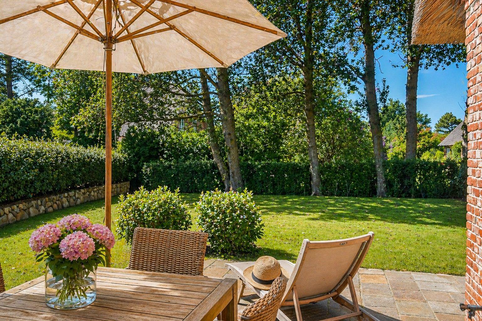 Terrasse mit Tisch, Stuhl und Sonnenschirm. Grünflächen und Bäume im Hintergrund.