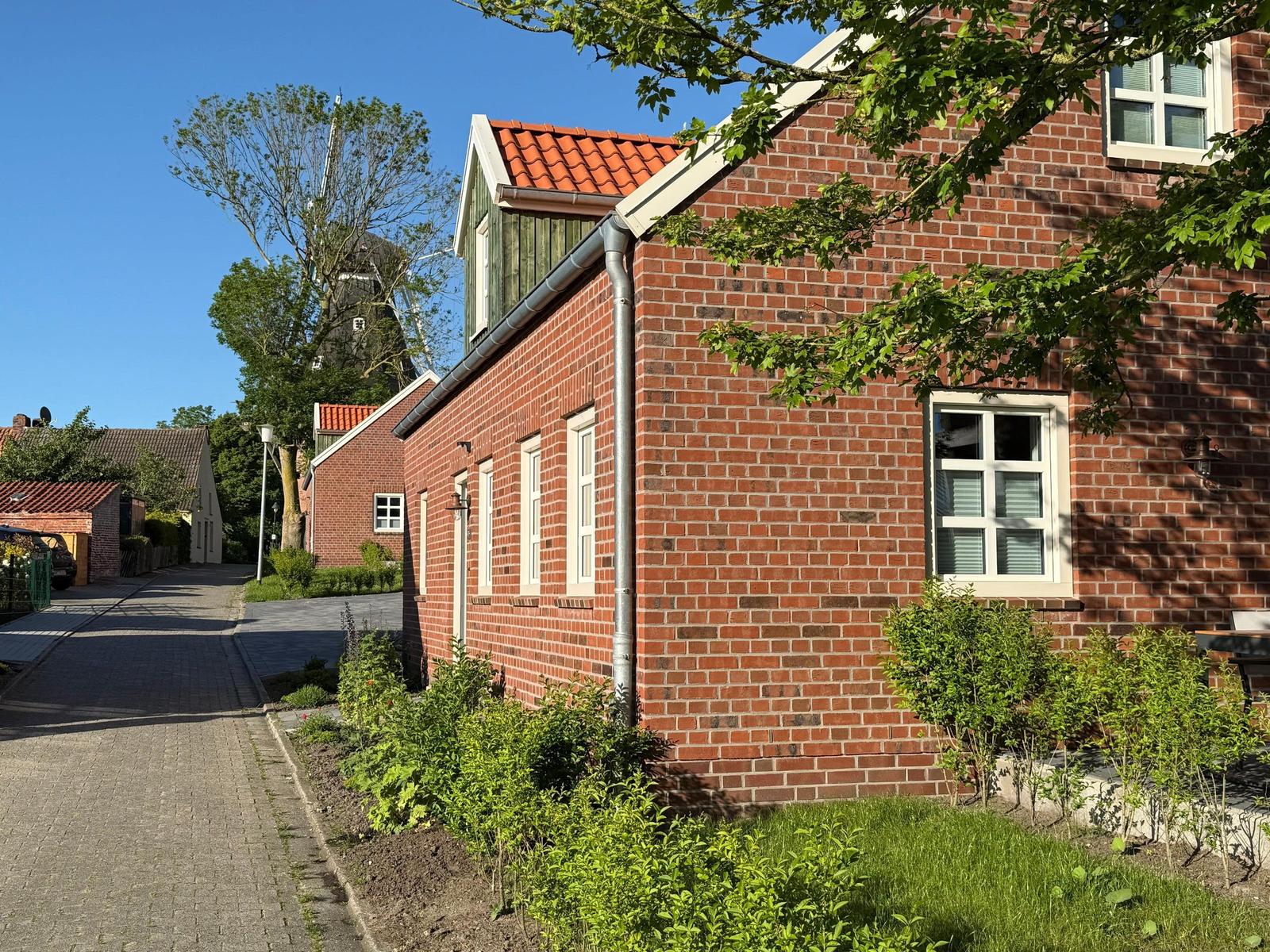 Rotes Backsteinhaus mit weißen Fenstern und rotem Dach. Grün bewachsenes Grundstück. Windmühle im Hintergrund.