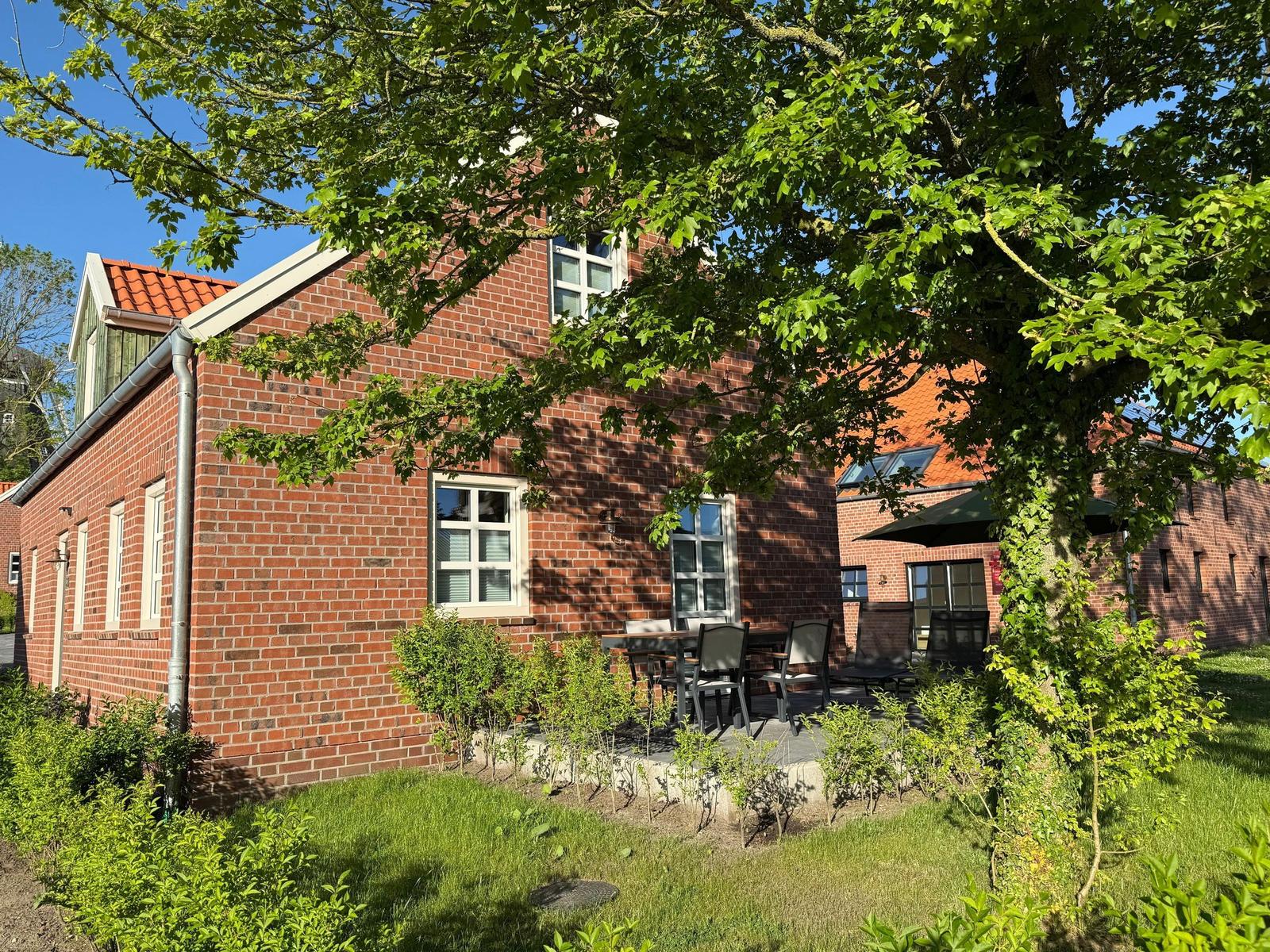 Rotes Backsteinhaus mit Terrasse, Garten und Bäumen unter blauem Himmel.