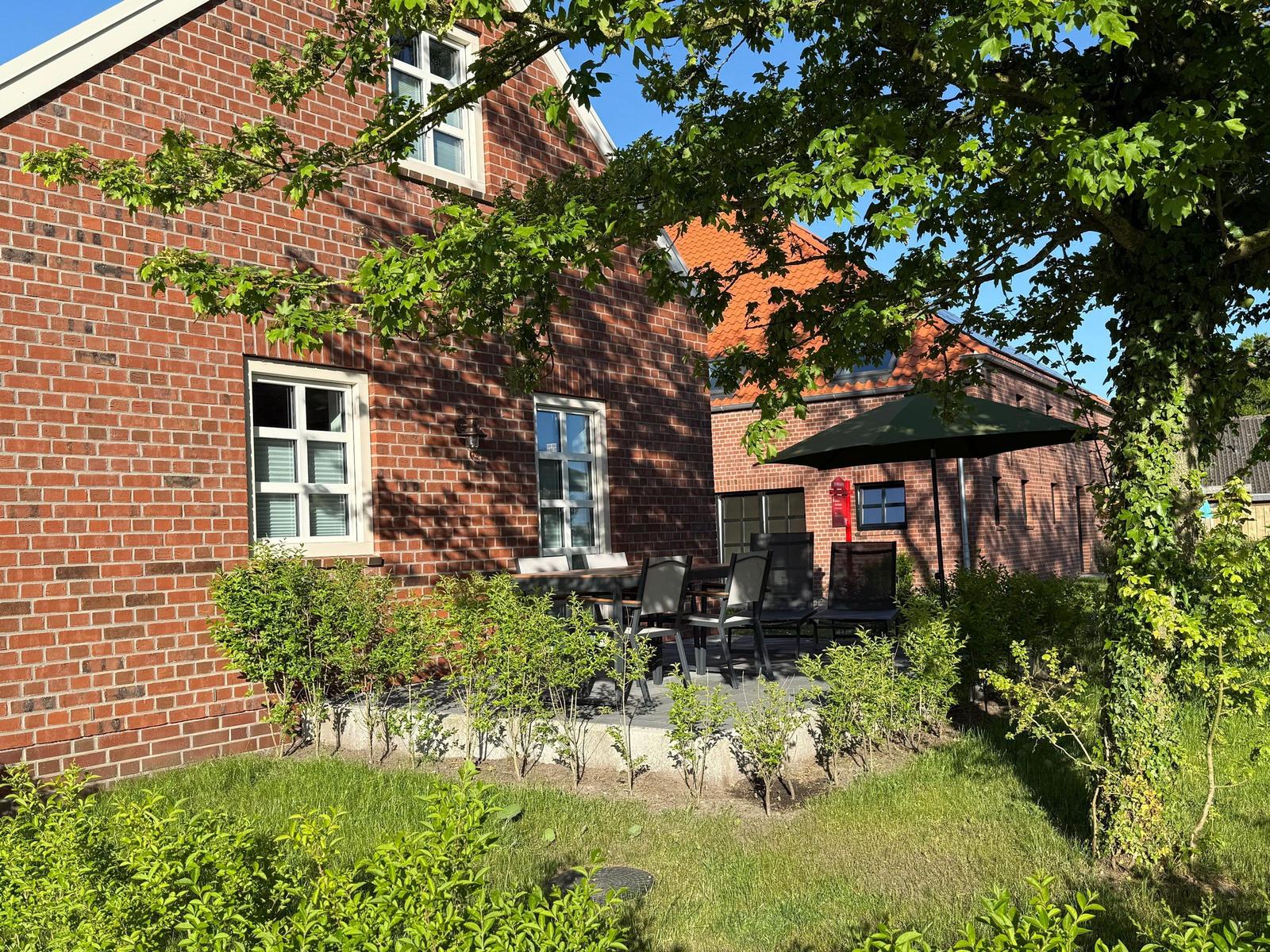 Rotes Backsteingebäude mit Terrasse, Tisch, Stühlen und Sonnenschirm unter Bäumen.