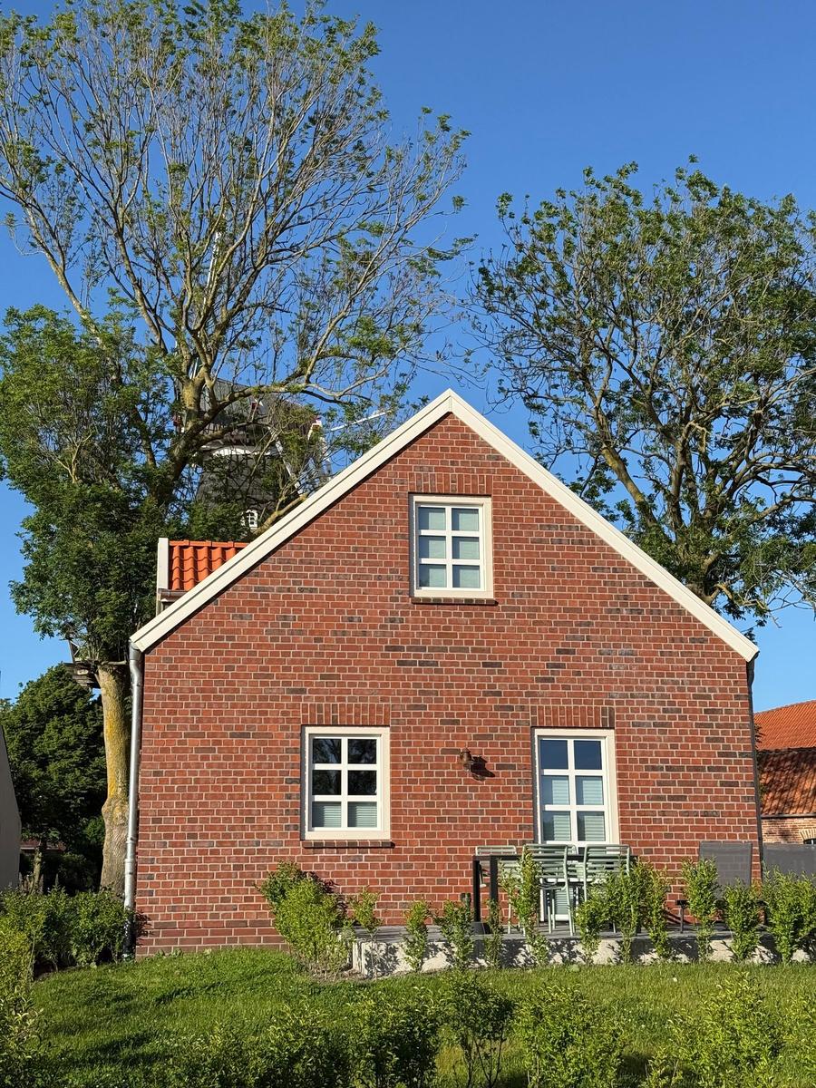 Rotes Backsteingebäude mit weißen Fenstern und Garten. Hintergrund: Bäume und klarer Himmel.