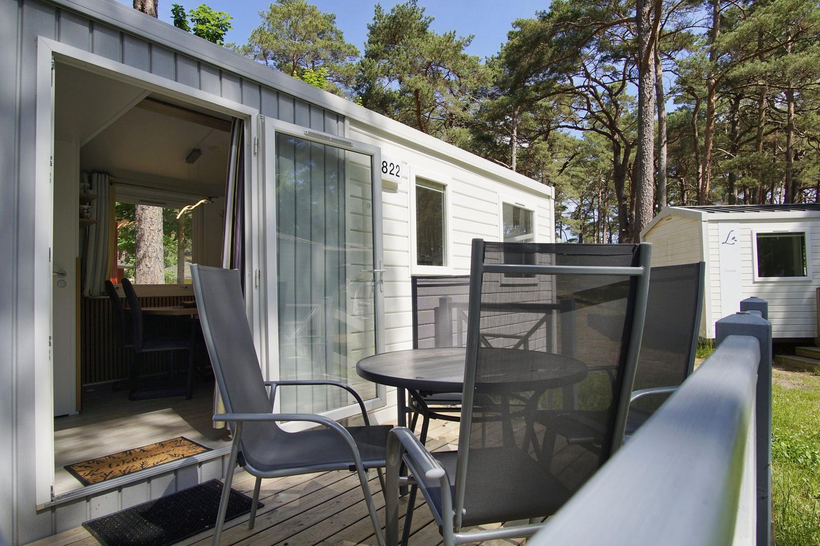 Terrasse mit Tisch und Stühlen vor einem weißen Ferienhaus mit Blick in den Wald.