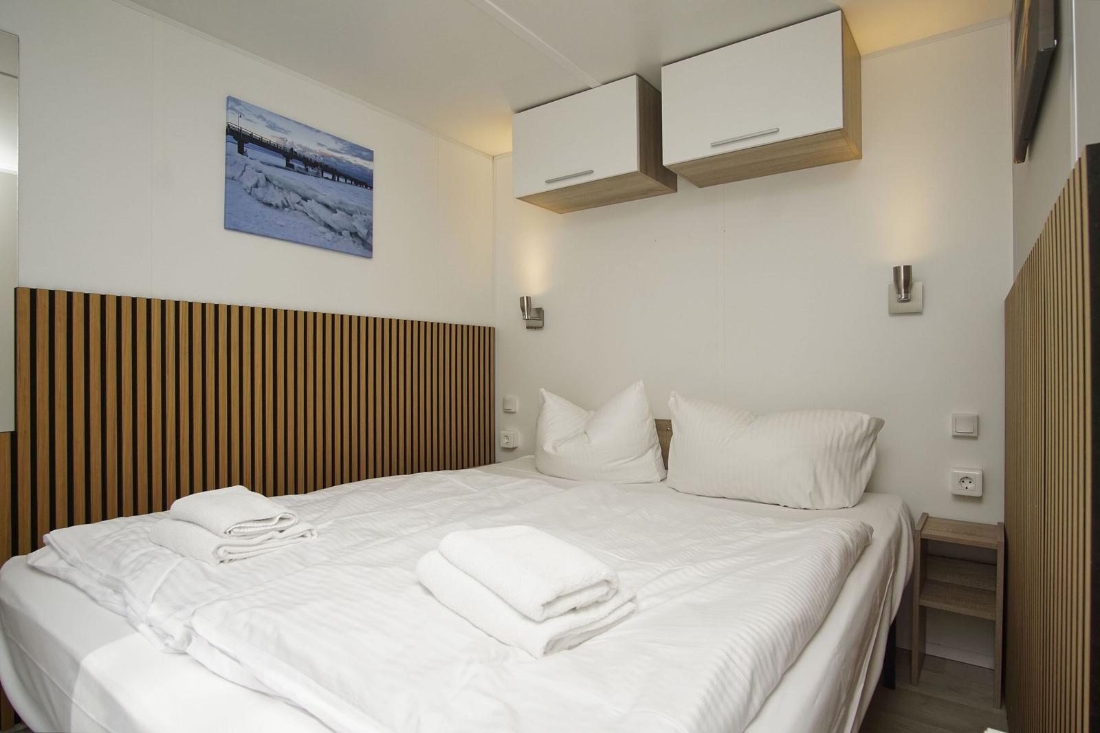 Schlafzimmer mit Bett, weißen Kissen und Handtüchern. An der Wand hängen Bilder und weiße Schrankkästen.