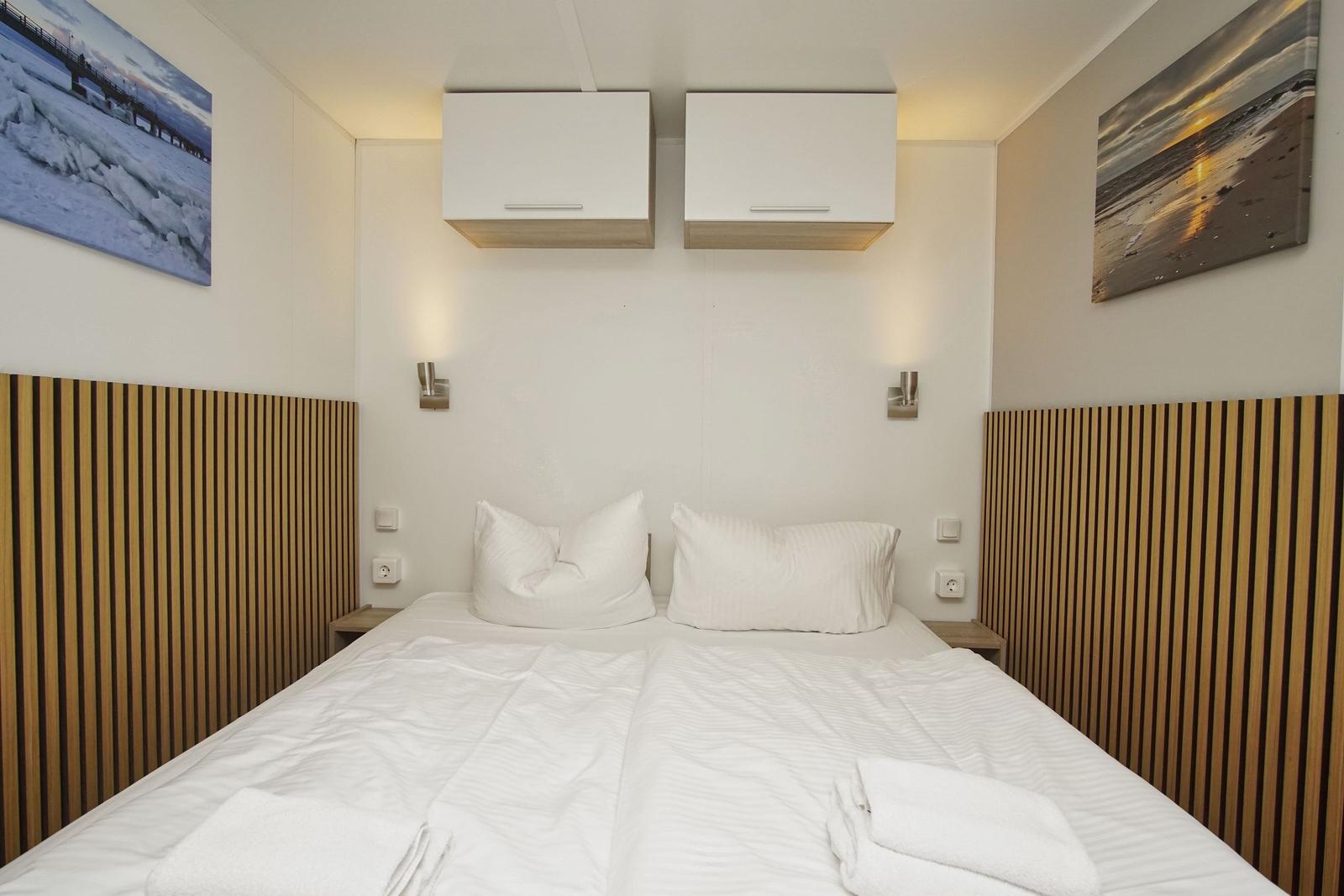 Schlafzimmer mit Doppelbett, weißen Bettwäsche, zwei Wandbildern und zwei Aufbewahrungskästen über dem Bett.