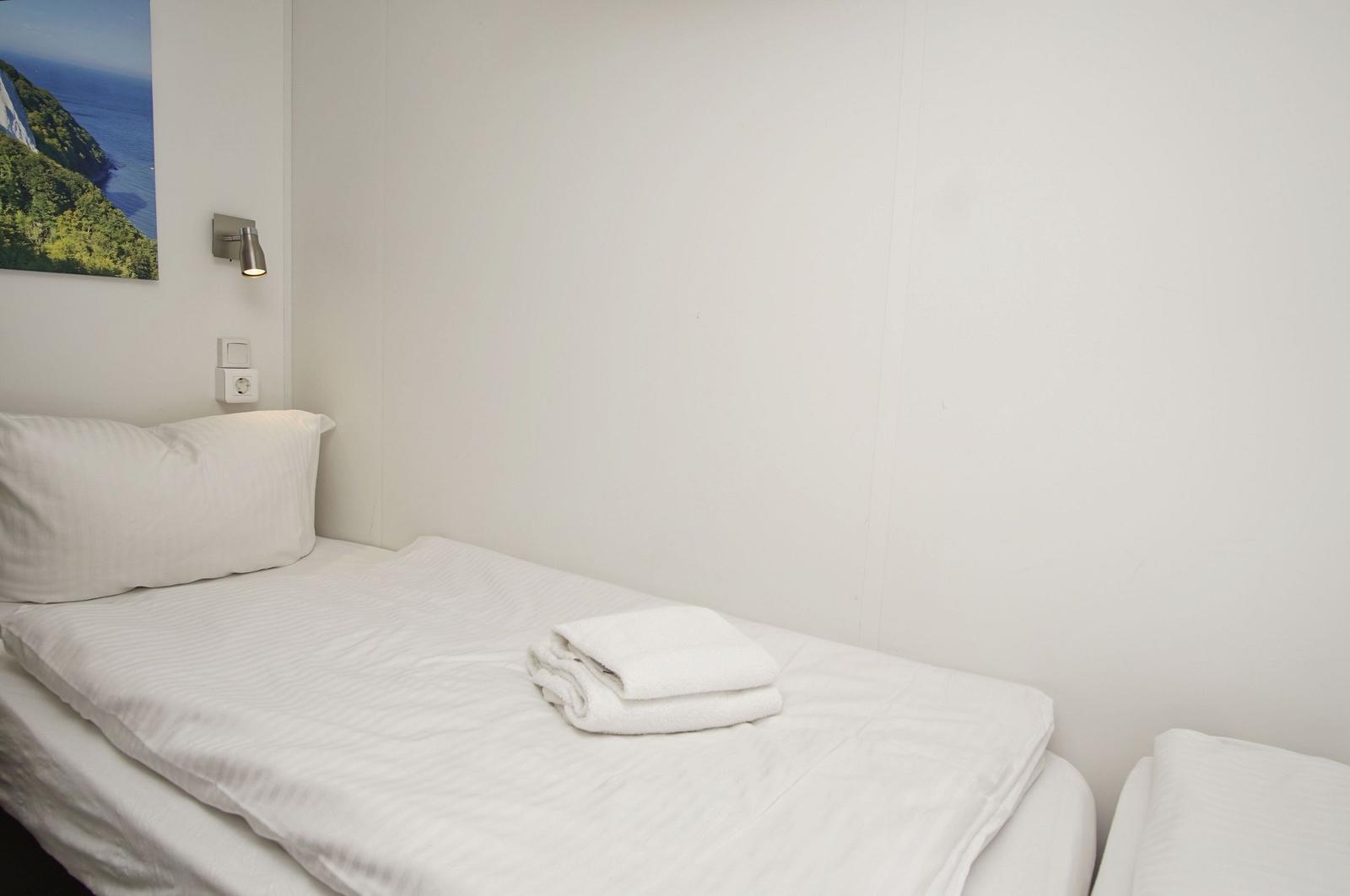 Ein Bett mit weißer Bettwäsche und einem gefalteten Handtuch. An der Wand hängt ein Bild und eine Wandlampe.