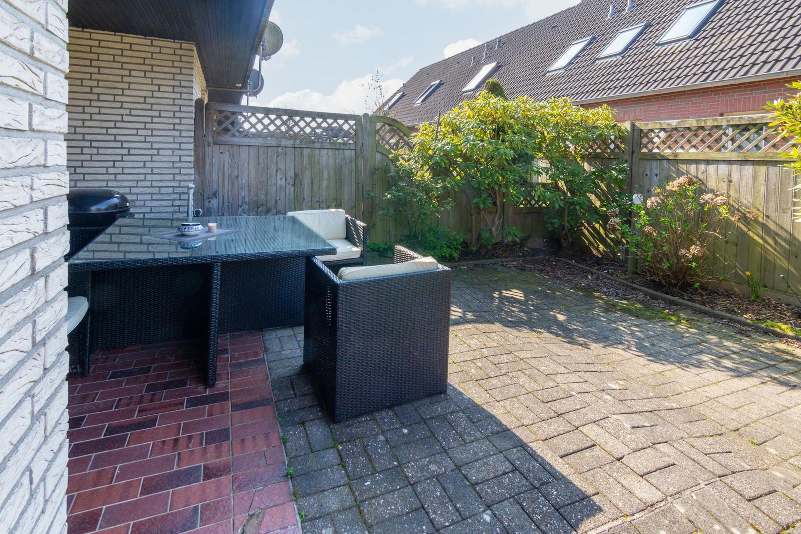 Terrasse mit Sitzgruppe, Grill und Pflanzen im Hintergrund.