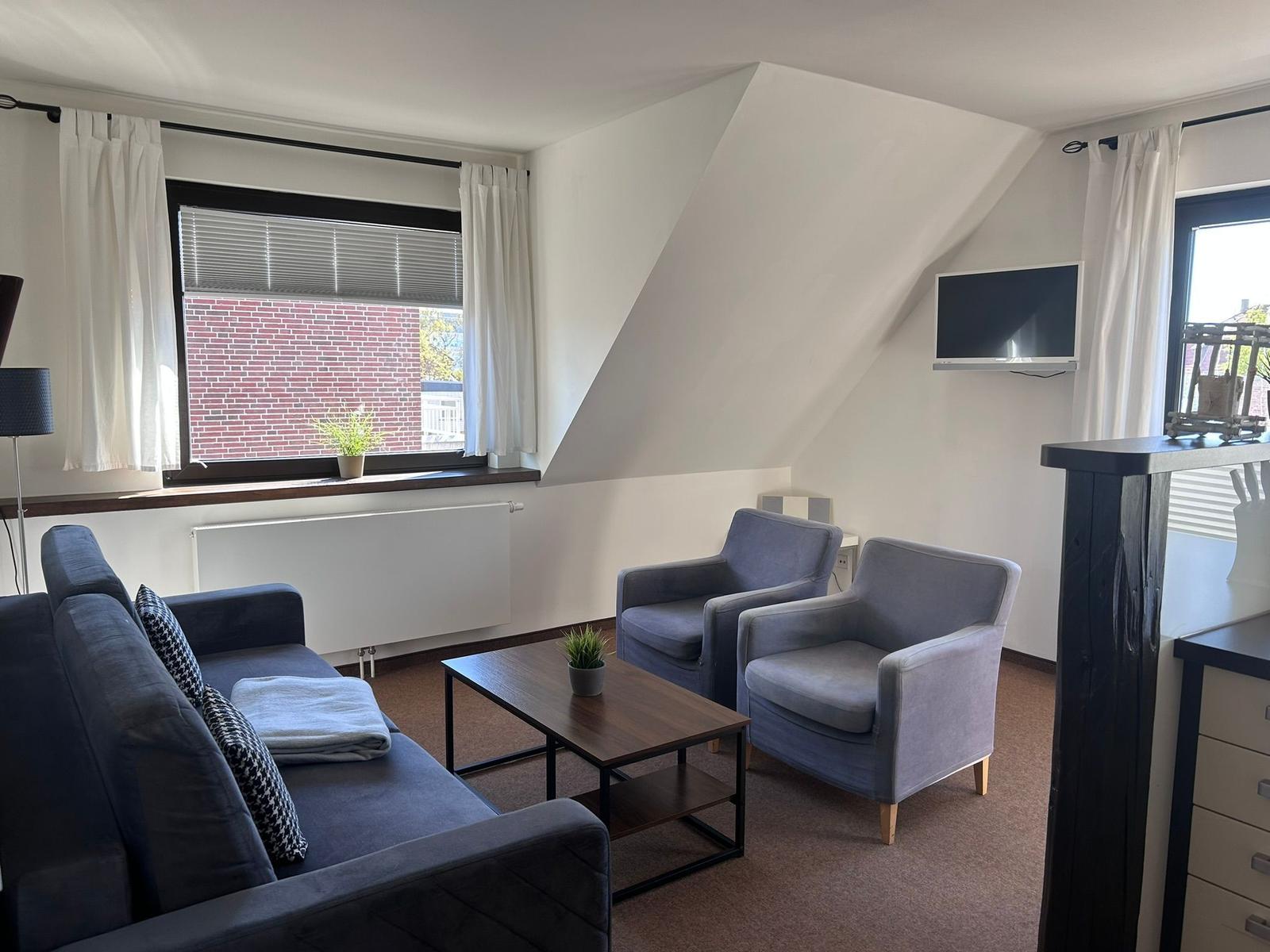 Wohnzimmer mit Sofa, zwei Sesseln, Couchtisch und Fernseher an der Wand.