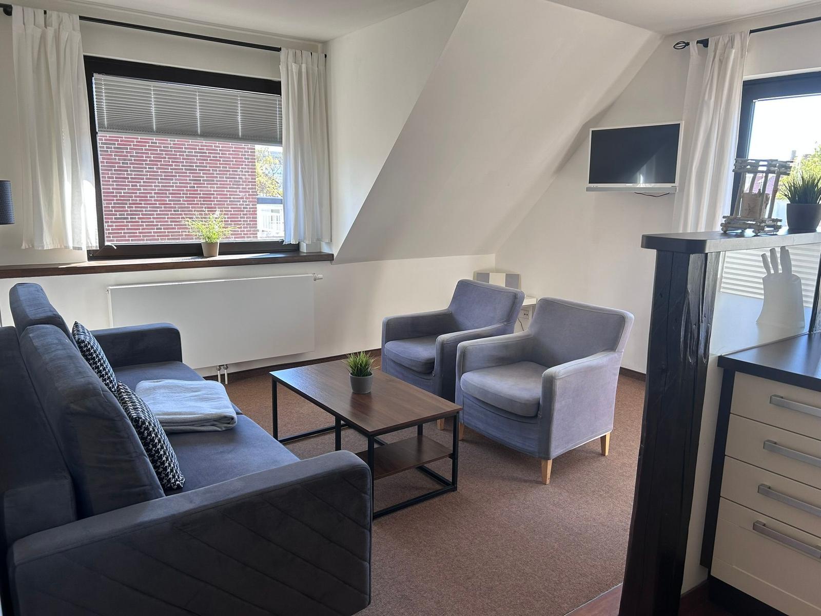 Wohnzimmer mit Sofa, zwei Sesseln, Couchtisch und Fernseher an der Wand.