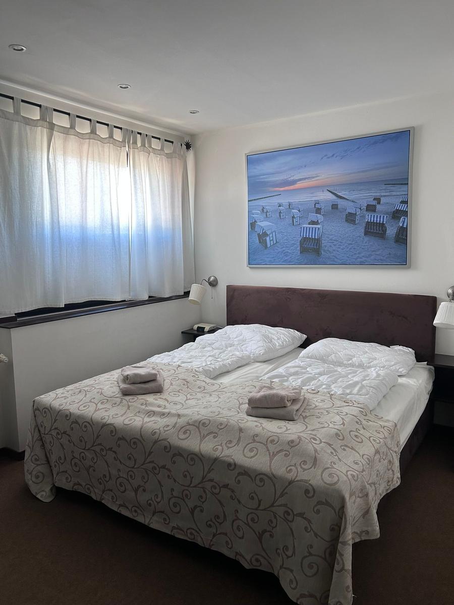 Doppelbett mit gemustertem Bettbezug, Wandbild mit Strandmotiv und Fenster mit weißen Vorhängen.