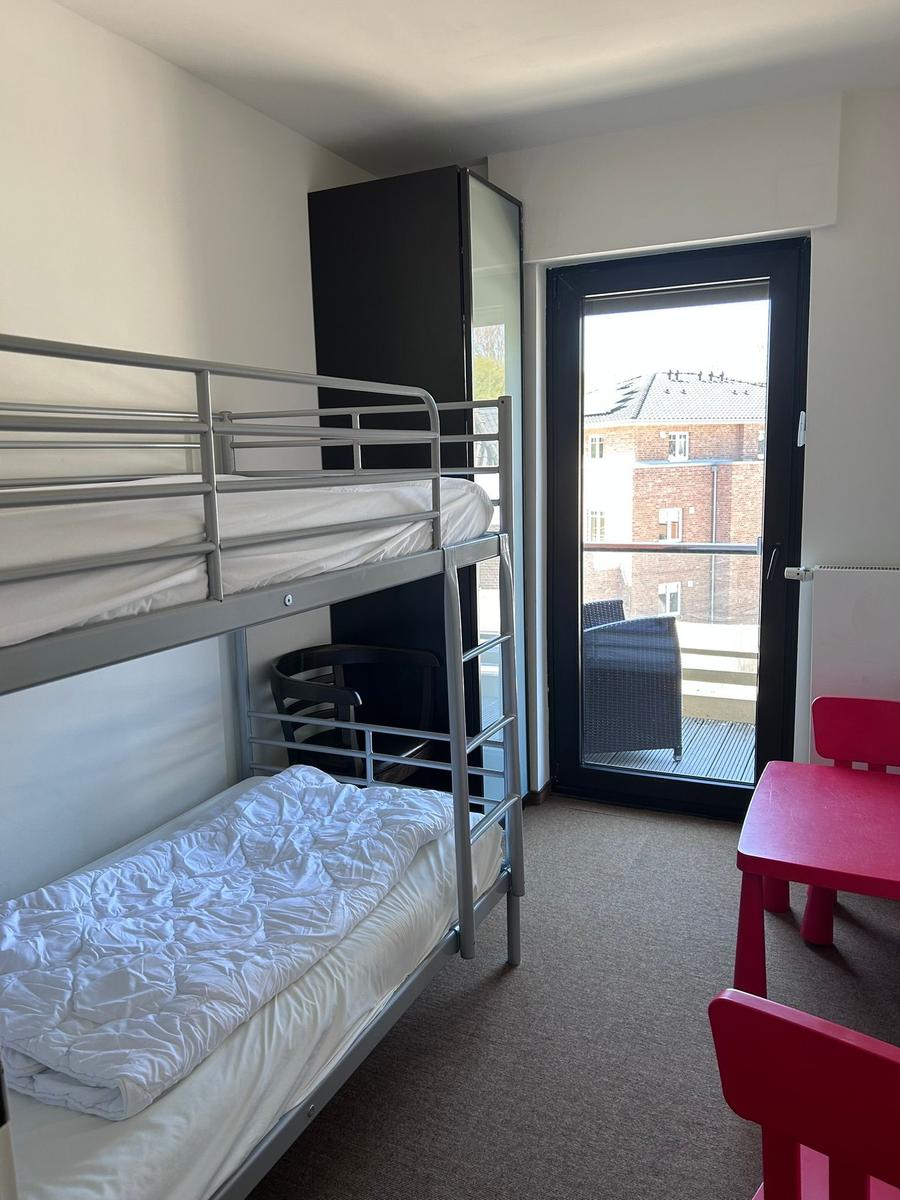 Zimmer mit Doppelstockbett, rotem Tisch und Stühlen, Balkon mit Blick auf Gebäude.