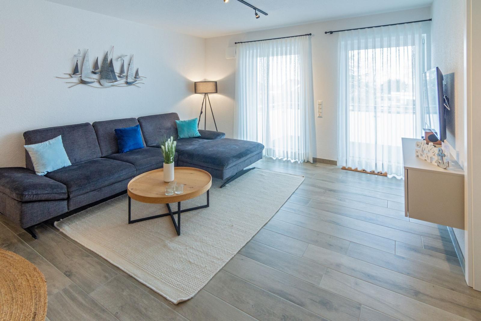 Ein Wohnzimmer mit grauem Sofa, runden Tisch und großen Fenstern.