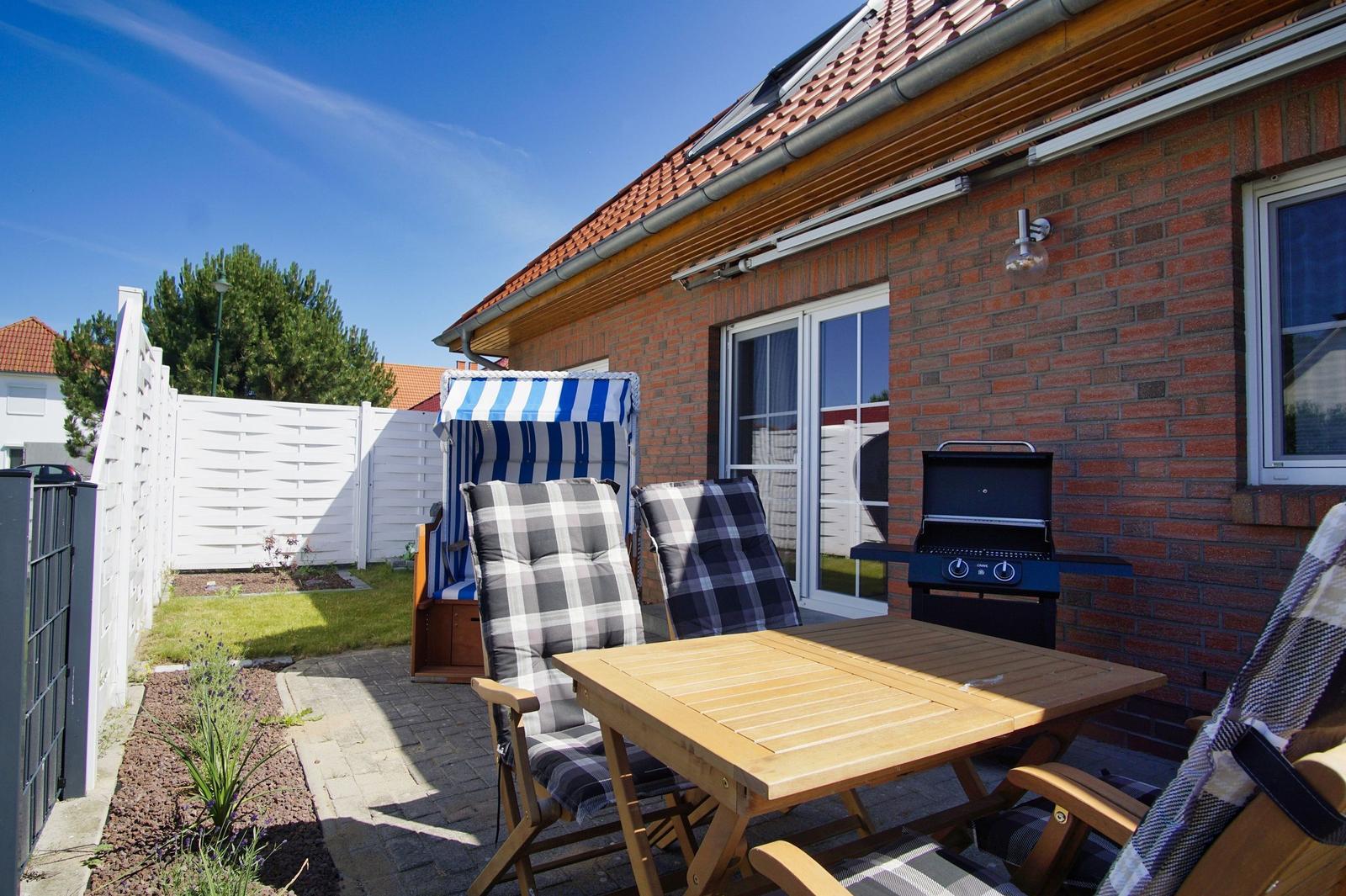 Terrasse mit Tisch, Stühlen, Grill und Strandkorb. Hinterhof mit Rasen und weißer Wand.