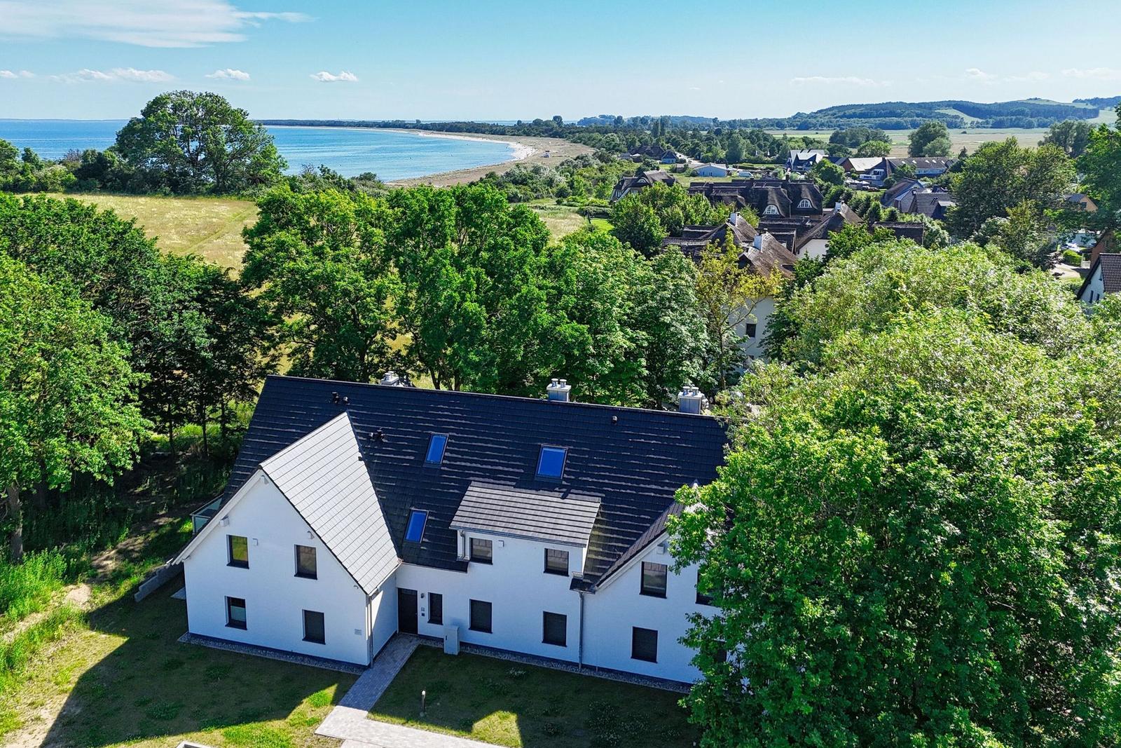 Modernes weißes Haus mit dunklem Dach, umgeben von Bäumen und Blick auf See und Strand.