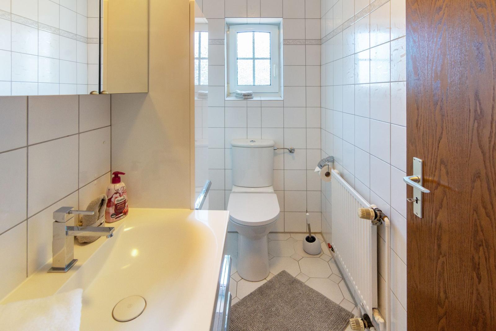 Badezimmer mit Waschbecken, Toilette und Fenster. Fliesenwand und Holztür.